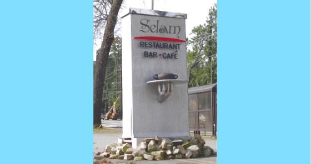 Sign for Selam Restaurant, a tall gray pillar with red text and a stone base, located outdoors.