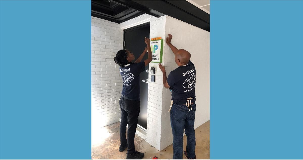 Two people installing equipment on a wall. One reaches up, installing a device. They wear matching blue shirts.