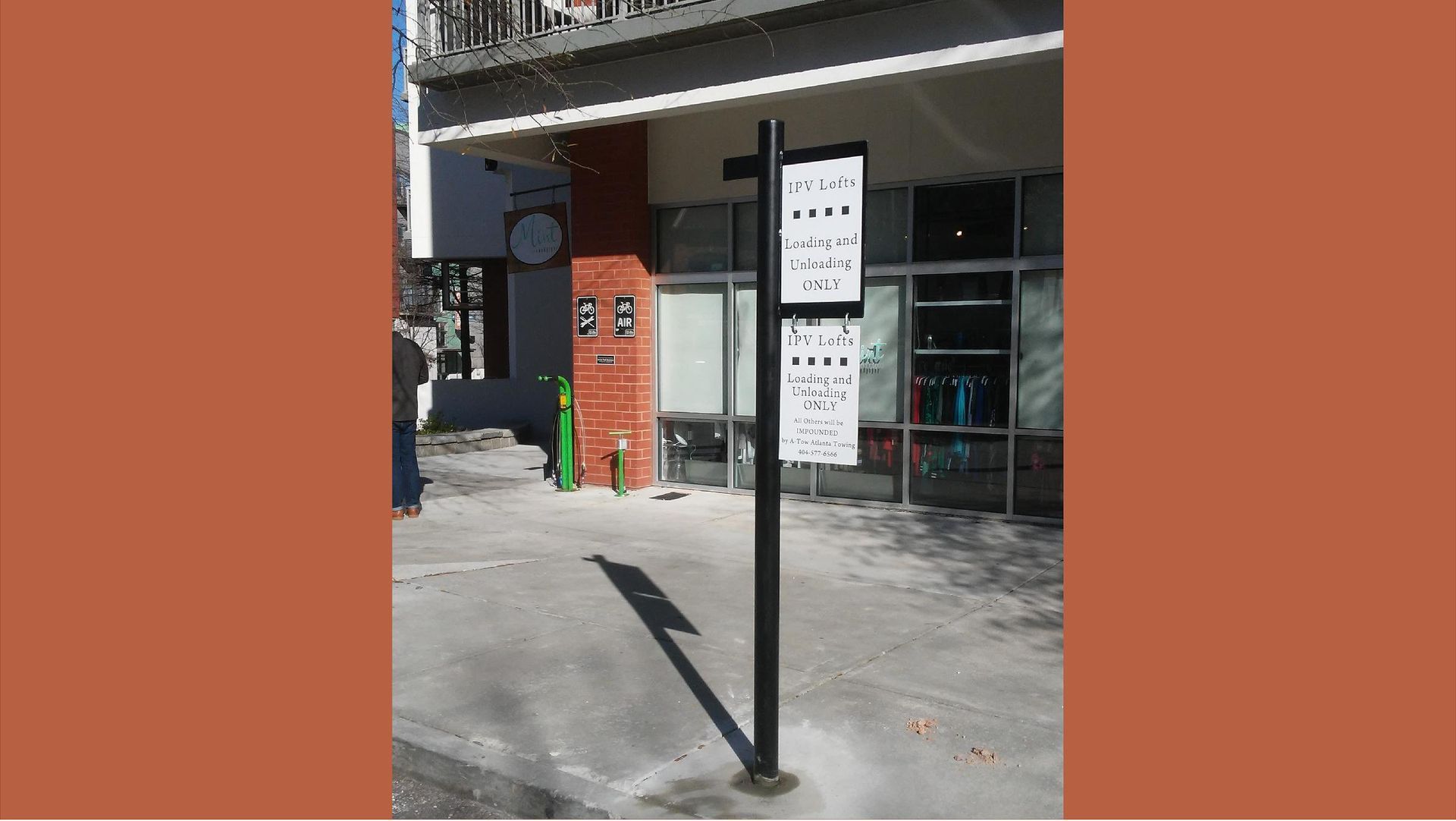 Black bus stop sign in front of a building with store windows. The sign is white and lists times.