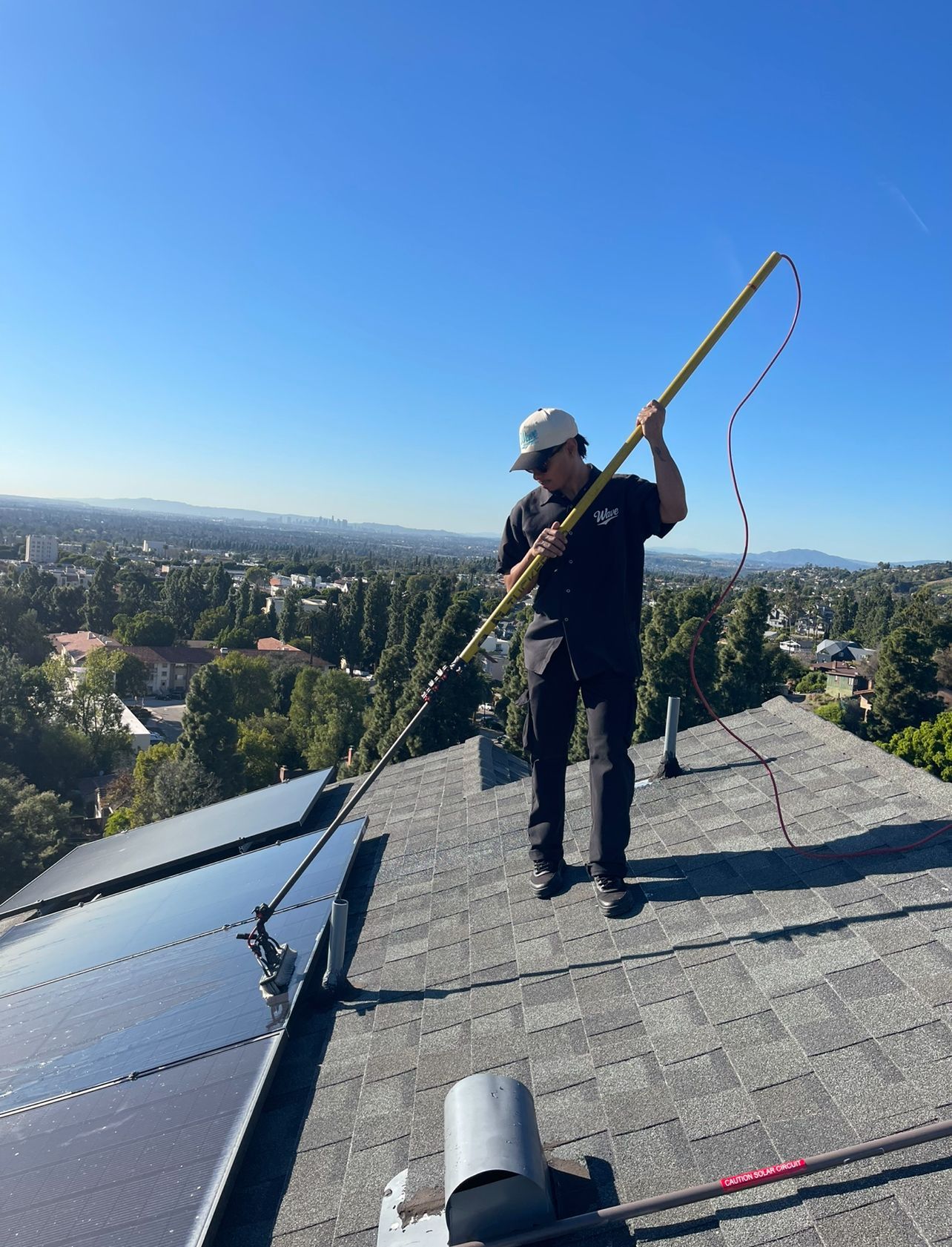 View from a roof showing a person using a yellow-handled tool on dark roofing panels and red tiles at sunset