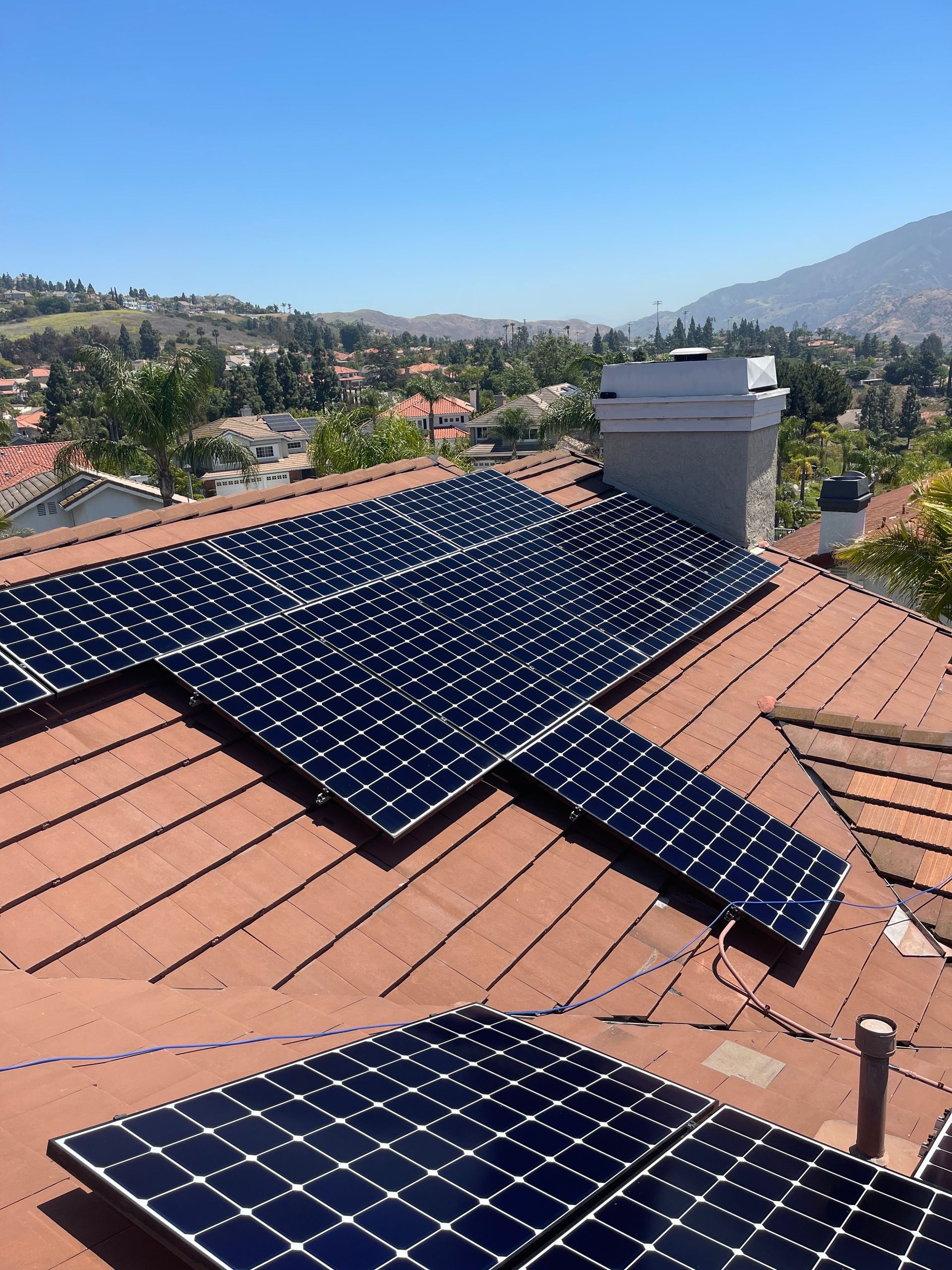 Solar panels on a terracotta roof overlooking a hilly suburban landscape under clear blue sky