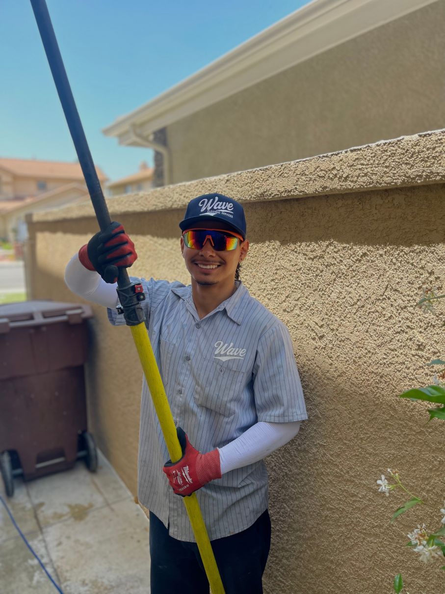 Person smiling and holding a long pole beside a stucco wall outdoors, wearing gloves and a cap.