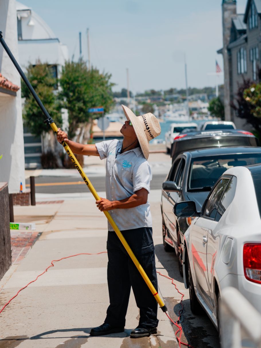 Worker in straw hat sprays a building exterior with a long pole sprayer on a sunny street.