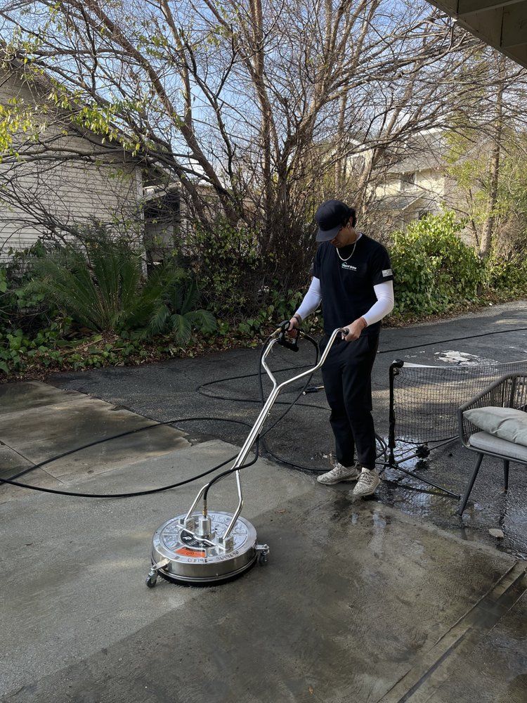 Person pressure-washes a wet concrete patio outdoors beside a house.