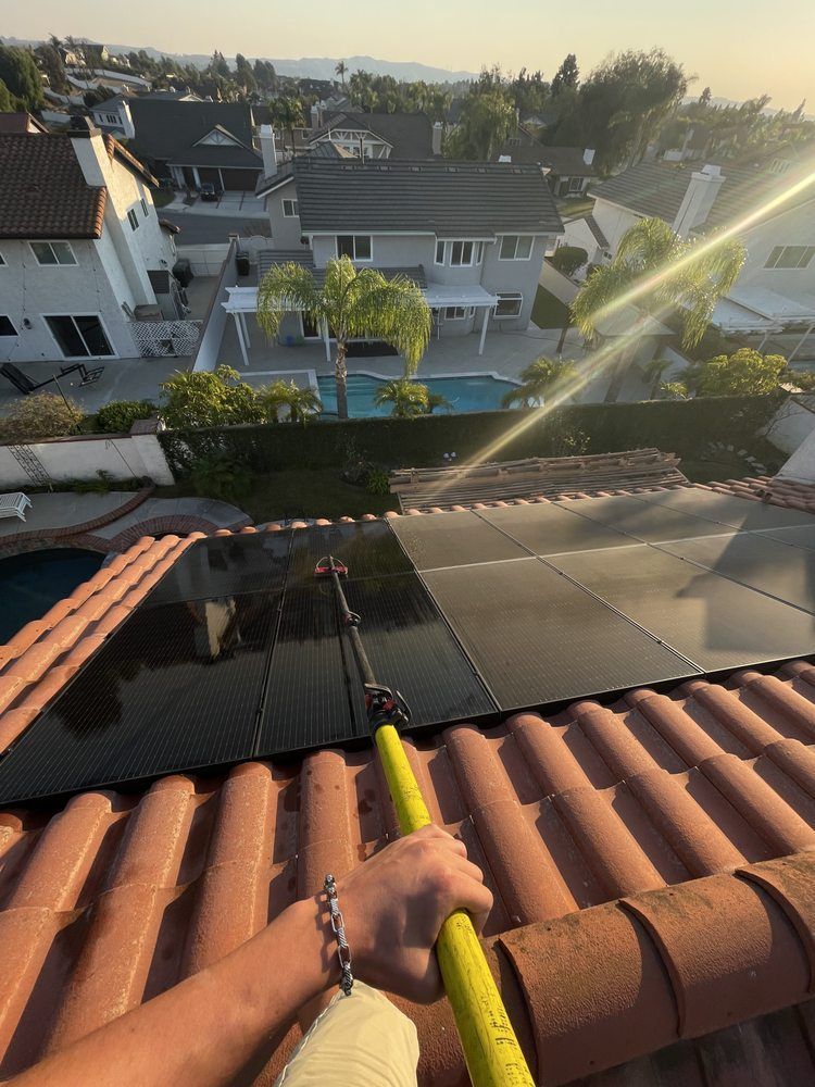 Hand holding a roof gutter tool above terracotta roof tiles, overlooking a suburban neighborhood at sunset