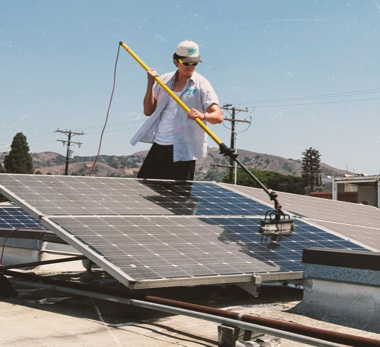 Technician cleaning rooftop solar panels with a long-handled brush under a clear sky