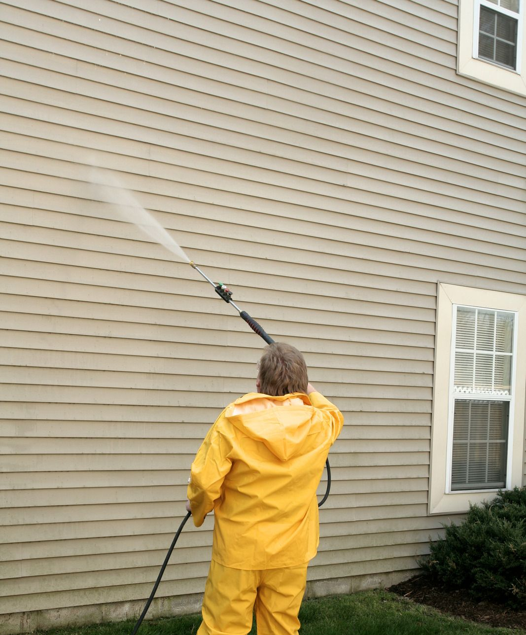 Person in yellow rain suit pressure-washing a house's siding.