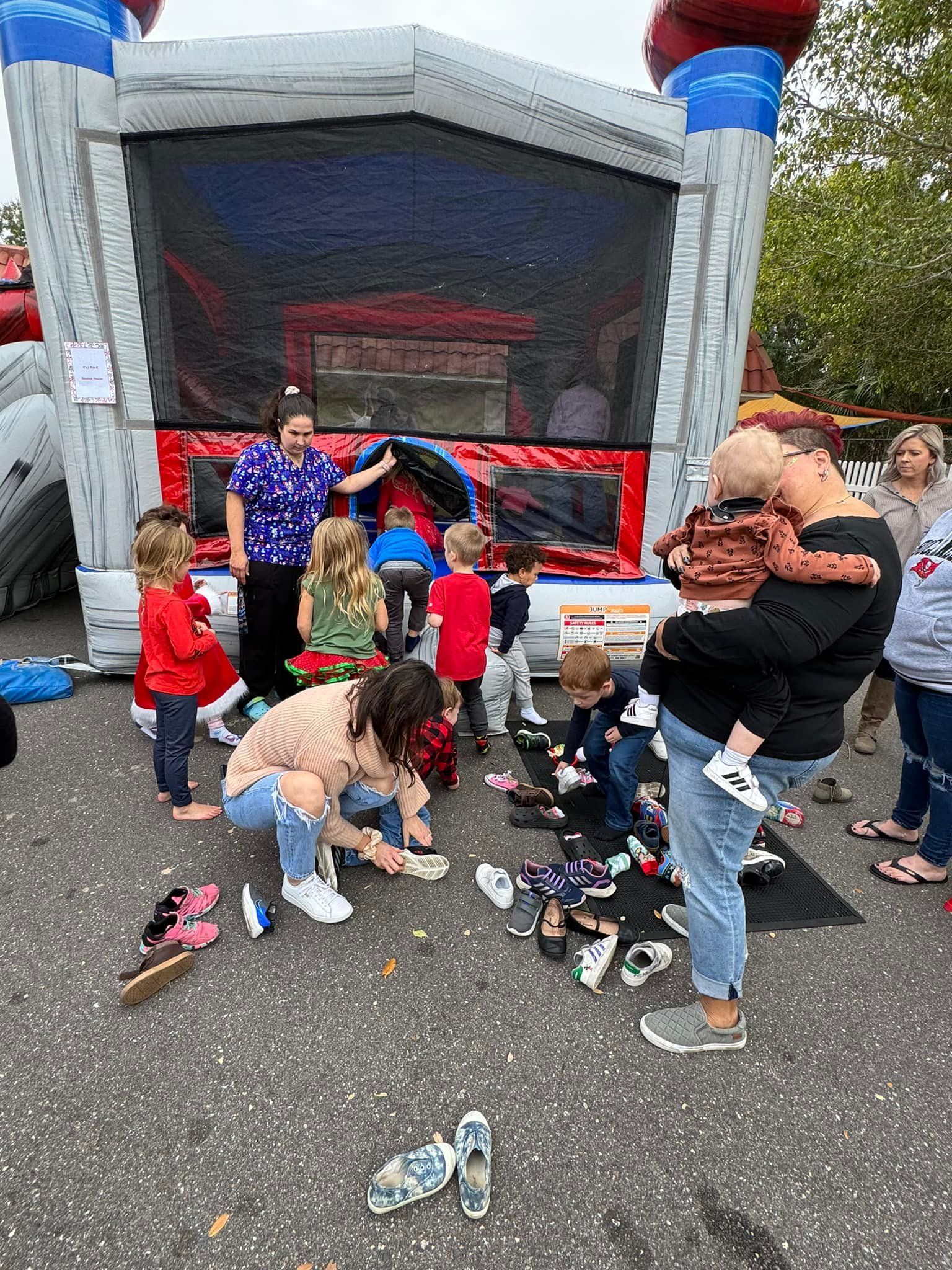 People gather outside a bounce house; children remove shoes while adults supervise.