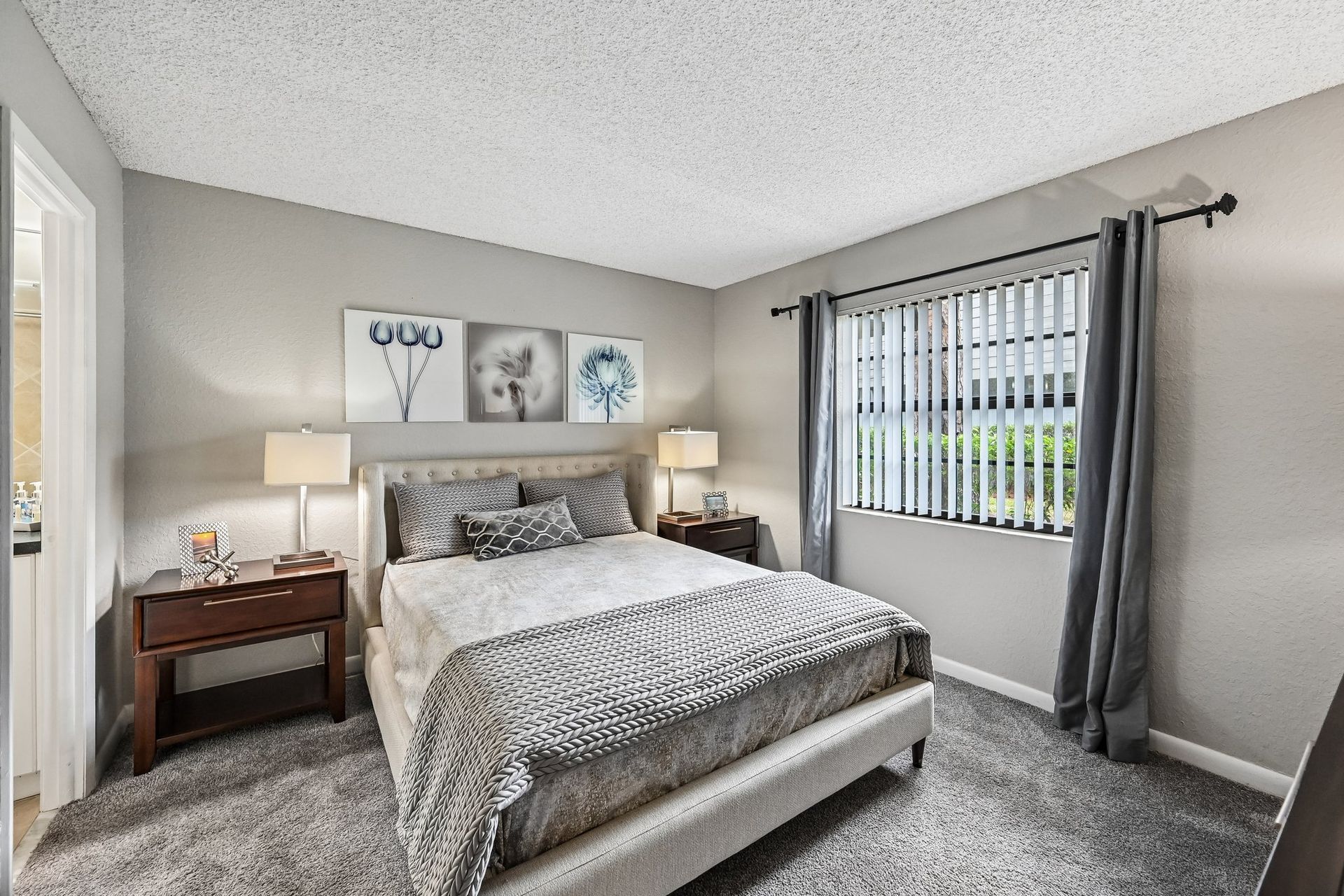 Bright gray bedroom with a queen bed, nightstands, wall art, and a window with curtains and blinds.