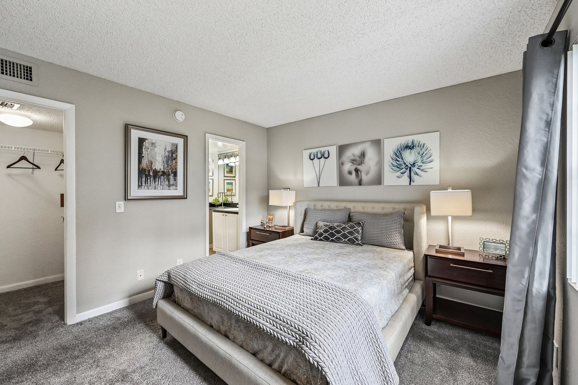 Bright bedroom with a made bed, gray walls, nightstands, lamps, and framed artwork above the headboard.