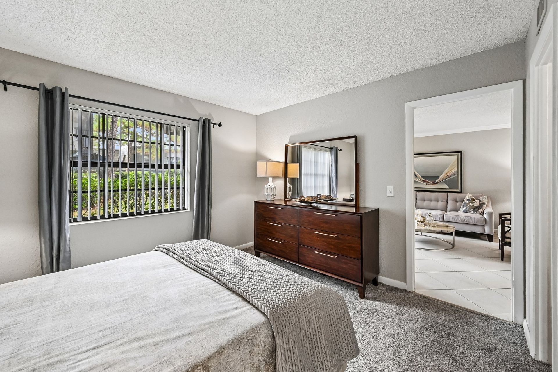 Bright bedroom with gray walls, bed, dresser with mirror, lamp, and window with curtains.
