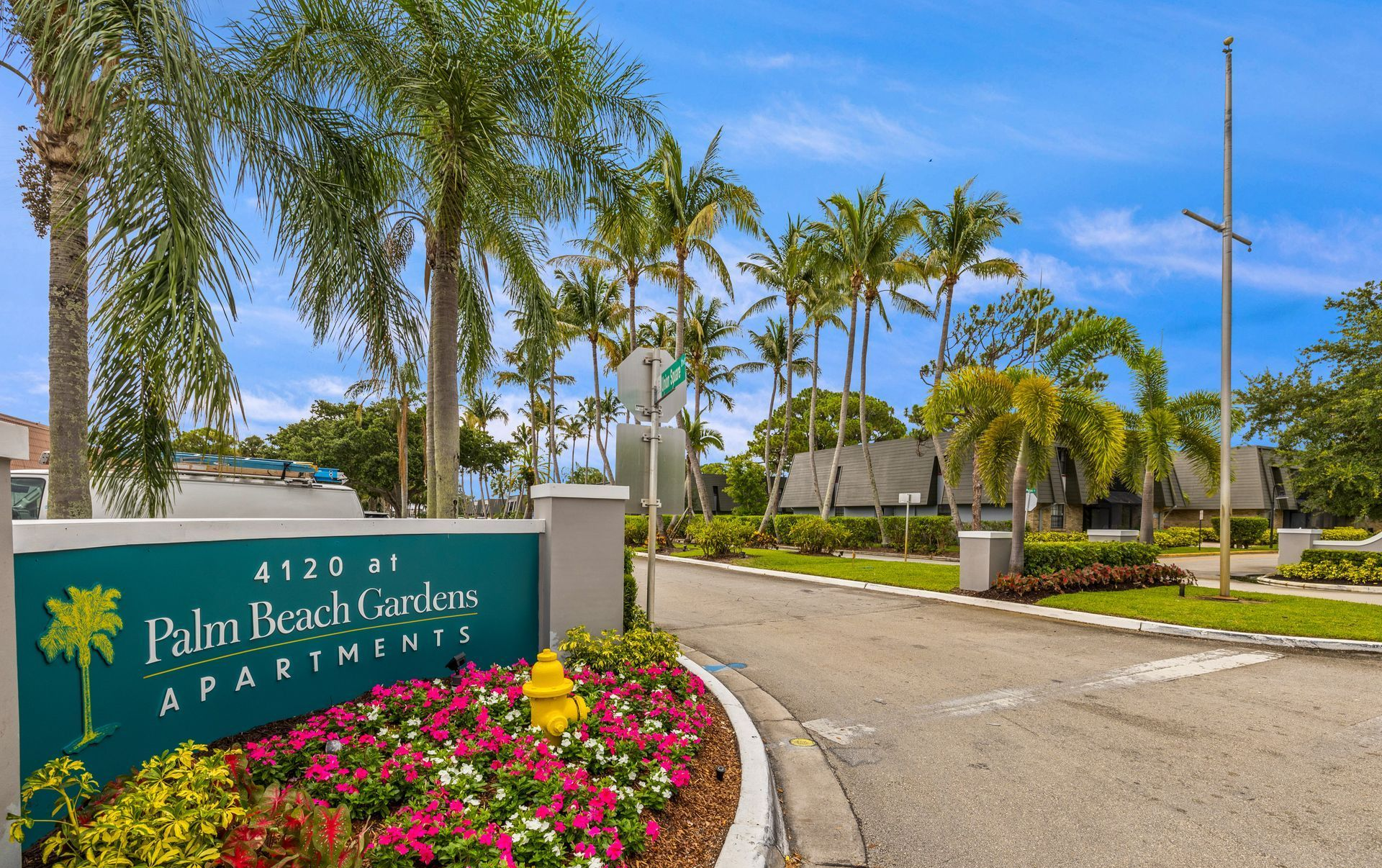 Entrance sign for Palm Beach Gardens Apartments beside a curving road, with palm trees and flowers.