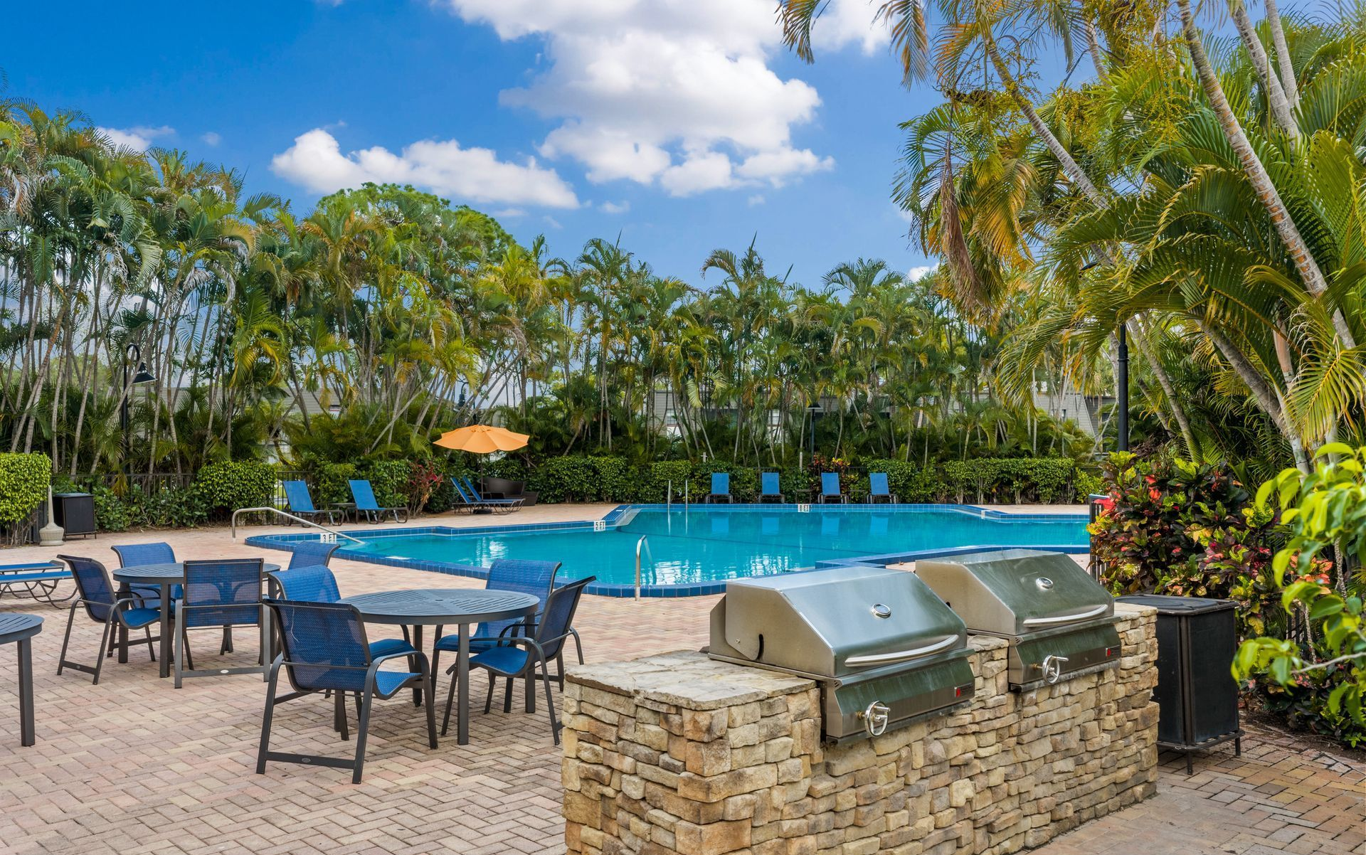 Poolside patio with tables and chairs beside a stone barbecue under palm trees and blue sky.