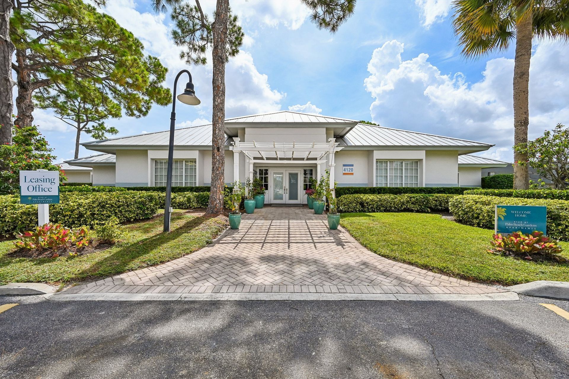 Front entrance of a white building with palm trees, hedges, and a paved walkway under a blue sky.