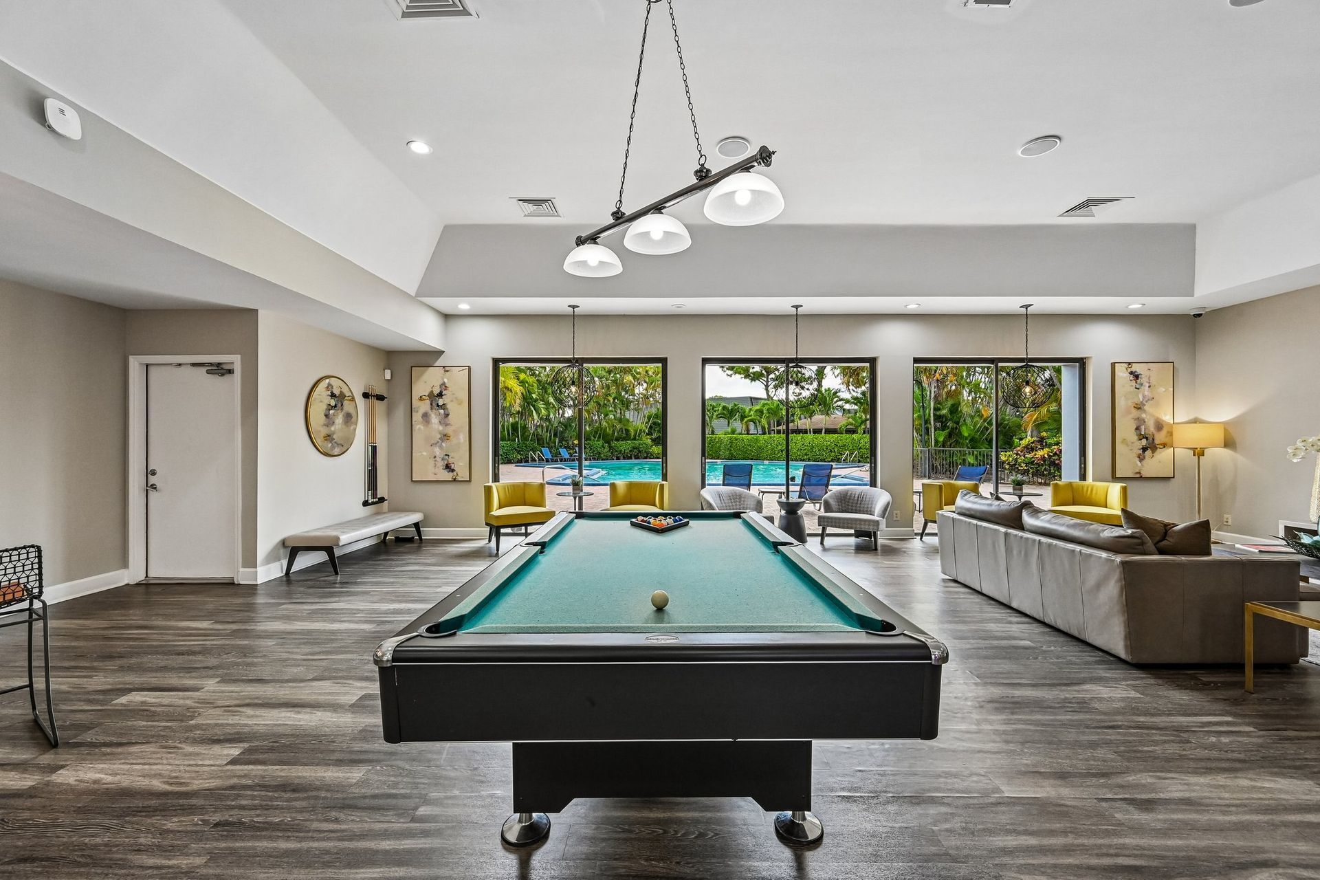 Modern game room with a pool table, gray sofa, and large windows overlooking a backyard pool.