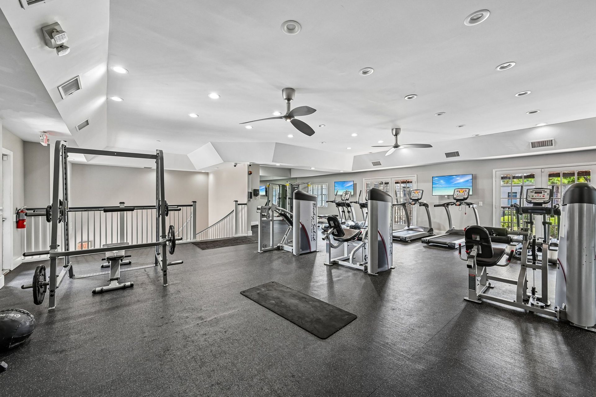 Bright gym with exercise machines, cable stations, and black rubber flooring under white ceilings.