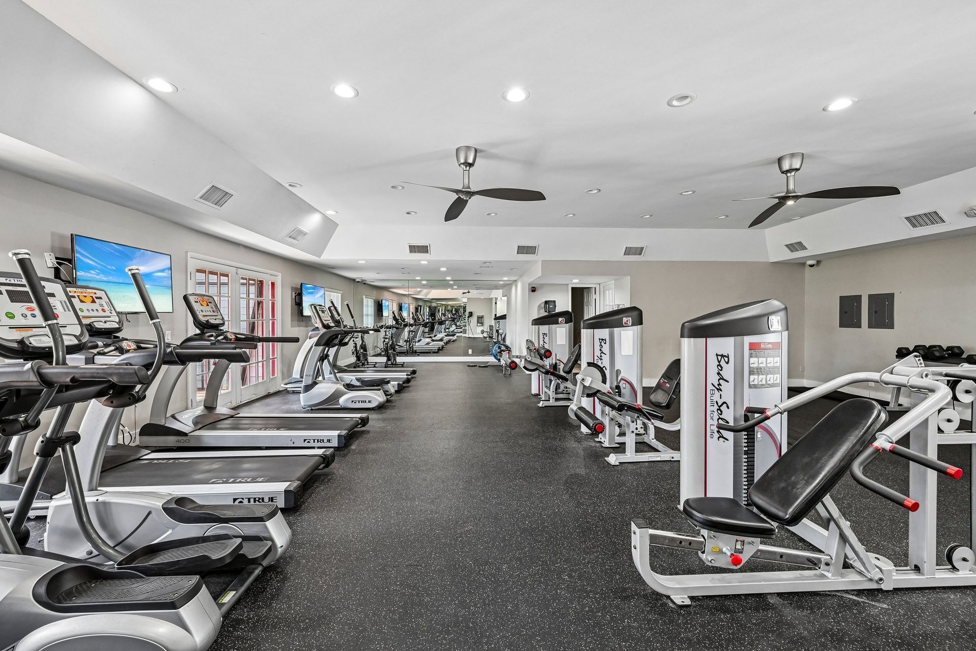 Bright, empty gym with treadmills, weight machines, and ceiling fans on a black rubber floor.