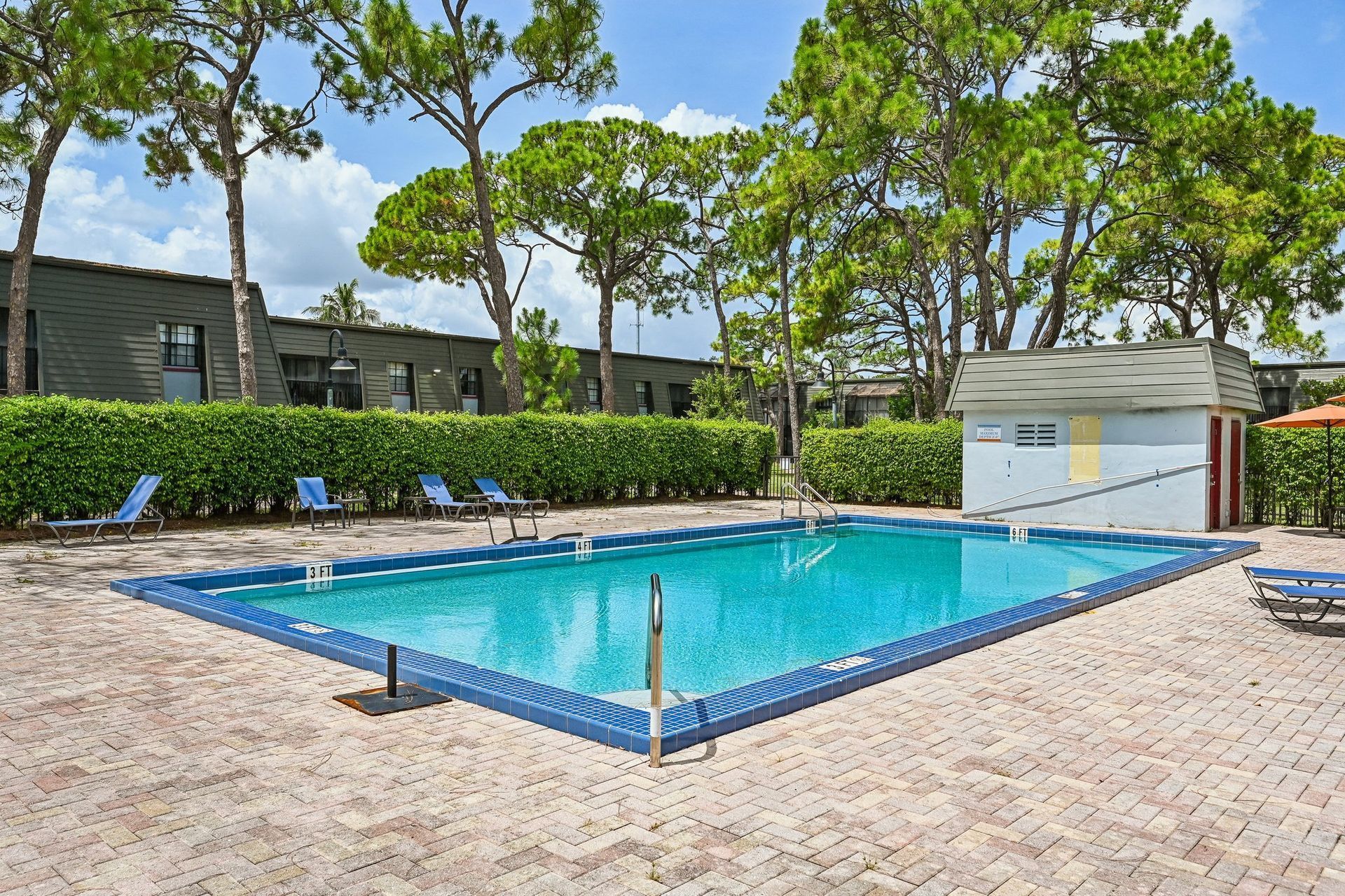Outdoor swimming pool with blue water, lounge chairs, trees, and a paved deck under a partly cloudy sky.