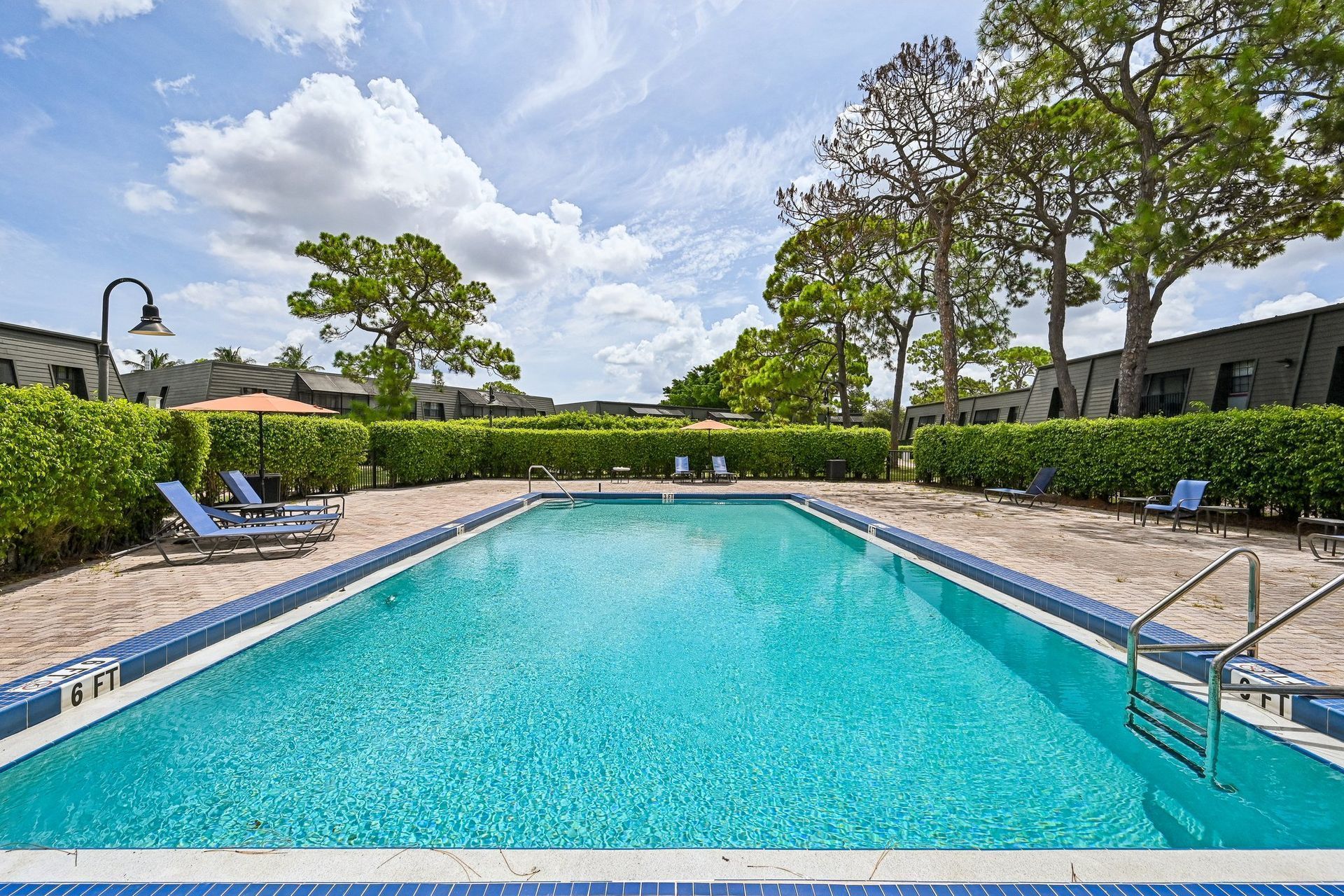 Outdoor swimming pool with turquoise water, lounge chairs, and trees under a partly cloudy sky.