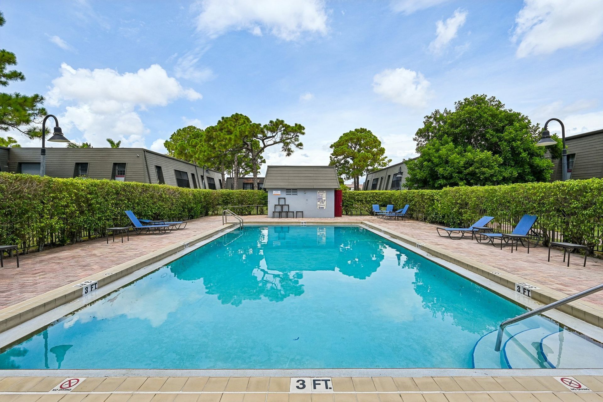 Outdoor swimming pool with turquoise water, lounge chairs, and hedges under a partly cloudy sky.