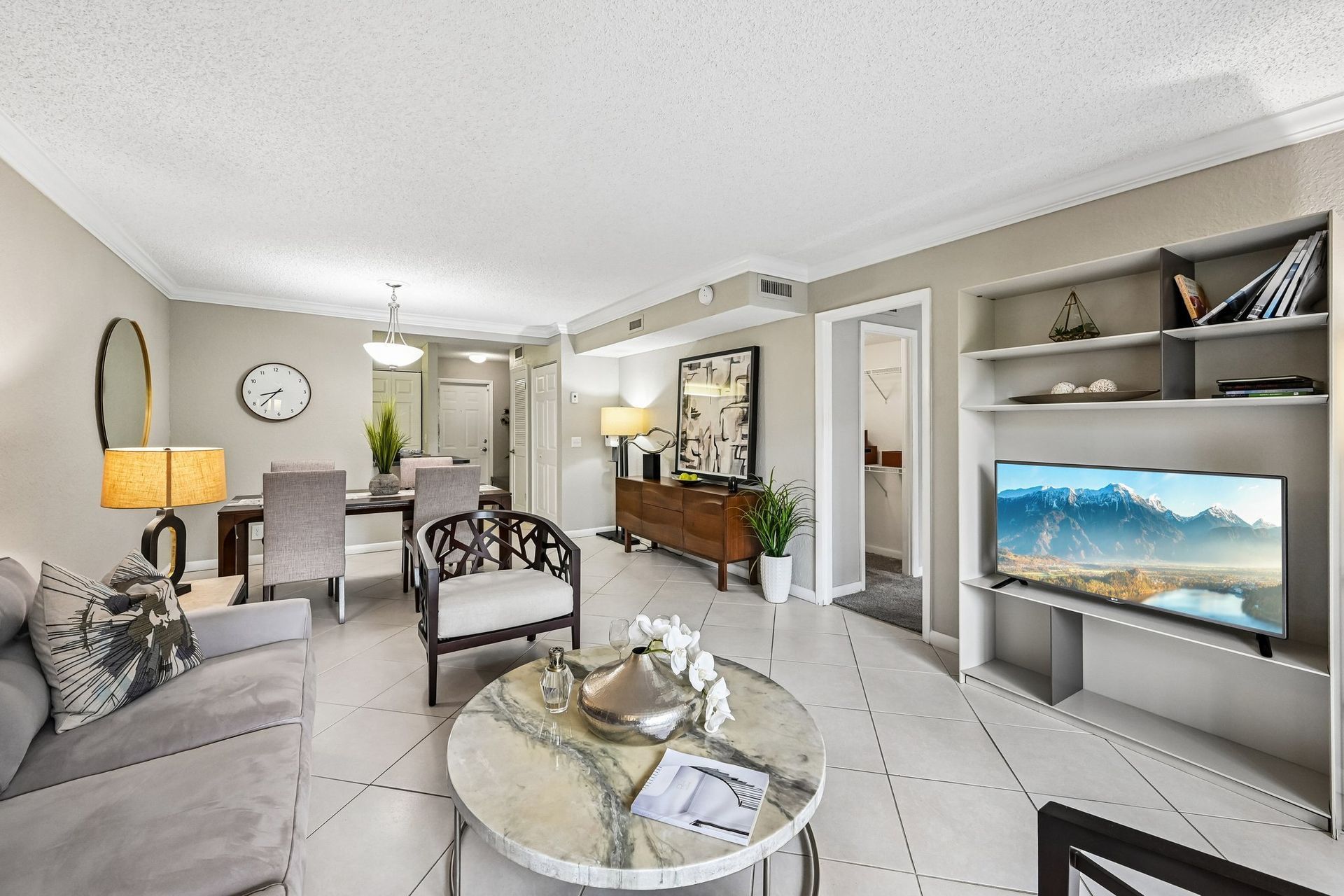Bright modern living room with gray sofa, TV, coffee table, dining area, and wall clock.