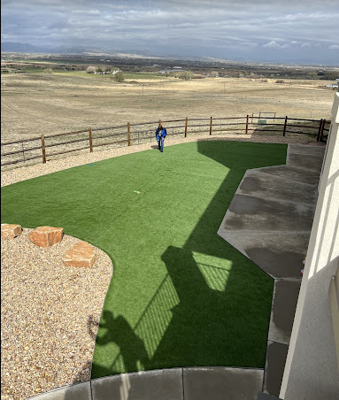 Backyard patio with artificial grass, gravel, fencing, and a person in blue standing near the edge.