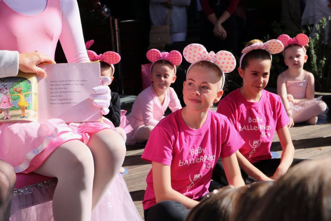 A Woman in a Pink Dress is Reading a Book to a Group of Young Girls — Studi-O Dance School In Gateshead, NSW