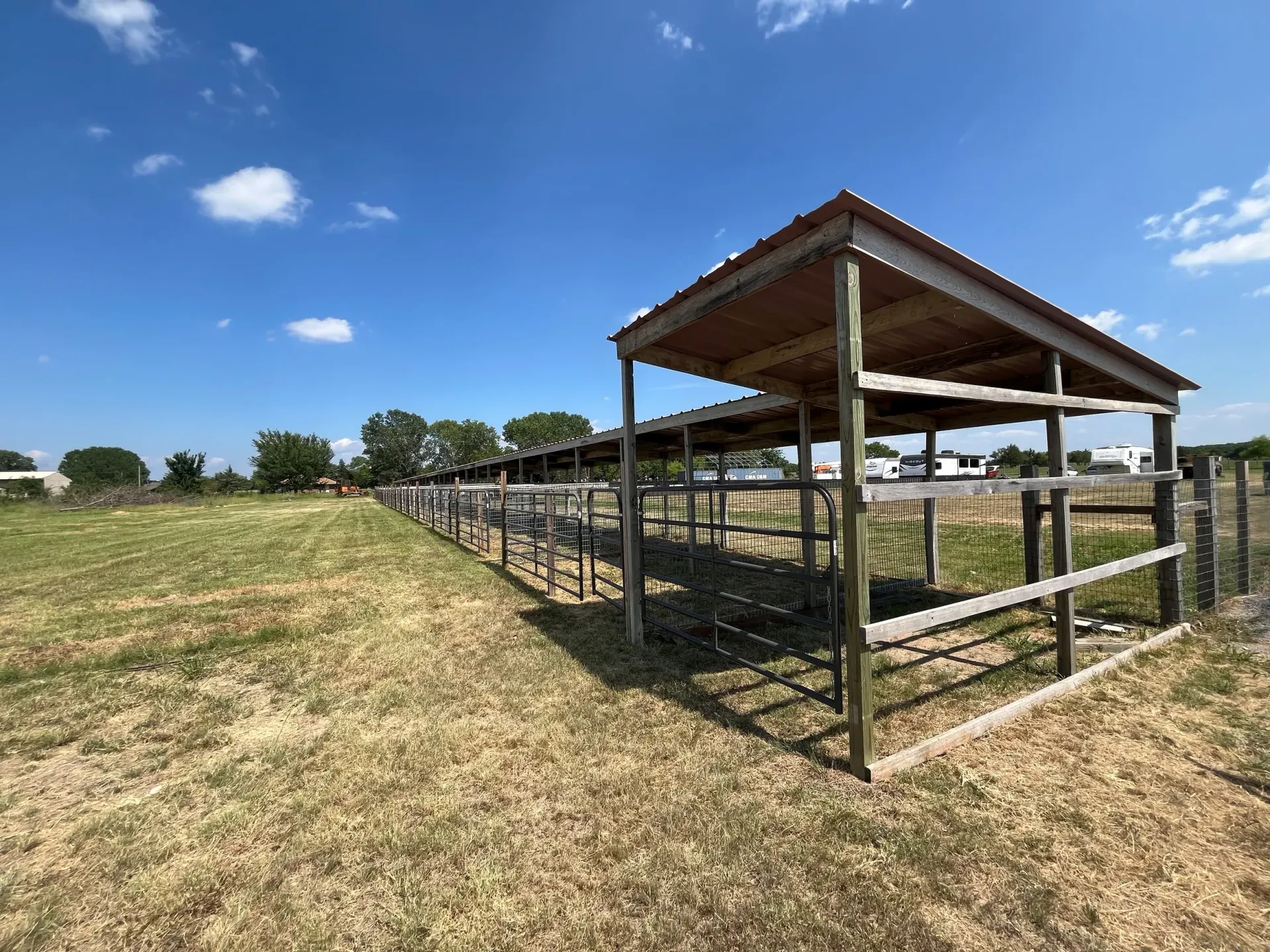 Open-sided livestock shelter with metal fencing on a grassy field under a blue sky.