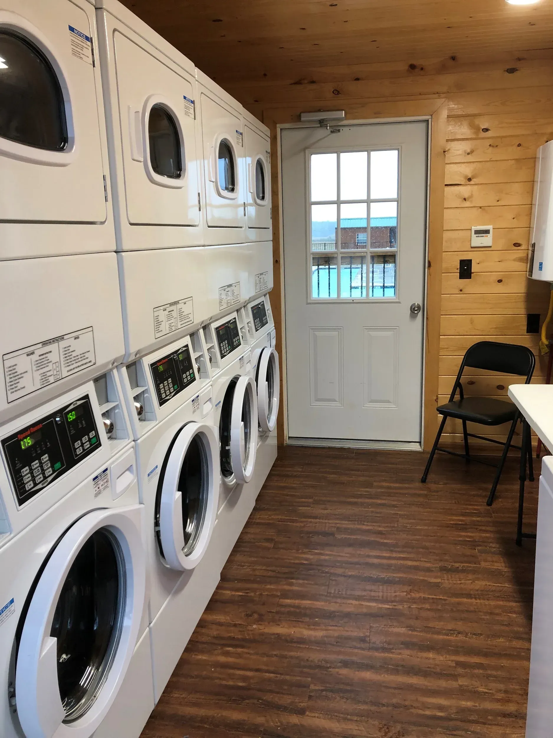 Laundry room with stacked washers and dryers, wooden walls and floor, and a white door at the end.