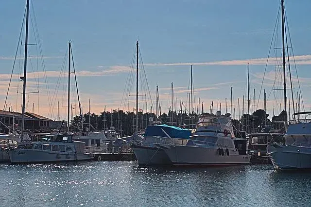 Boats docked in a marina at sunset, with calm water and tall masts silhouetted against a pastel sky.