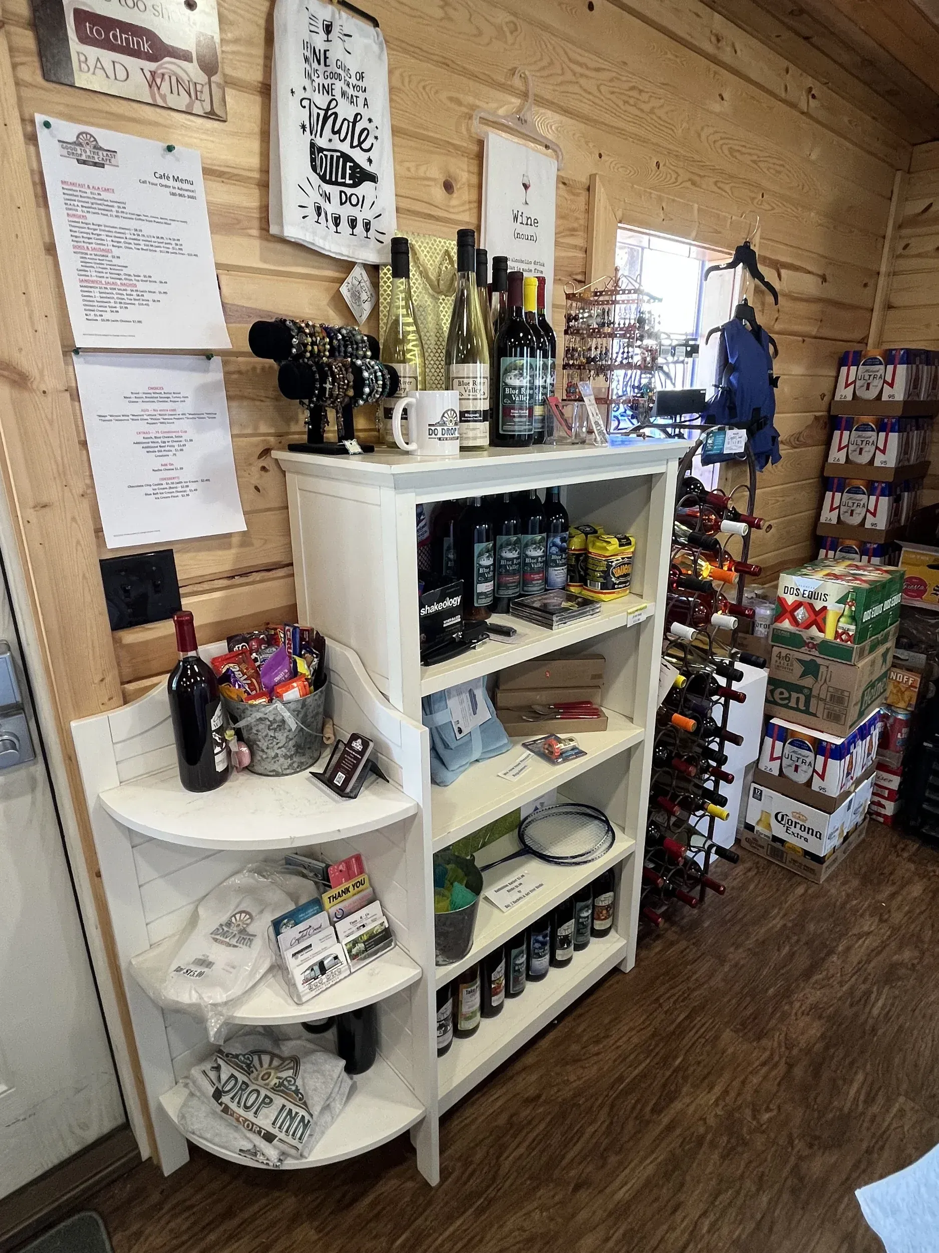Small shop display with shelves of wine bottles, snacks, and hanging menus against a wood wall