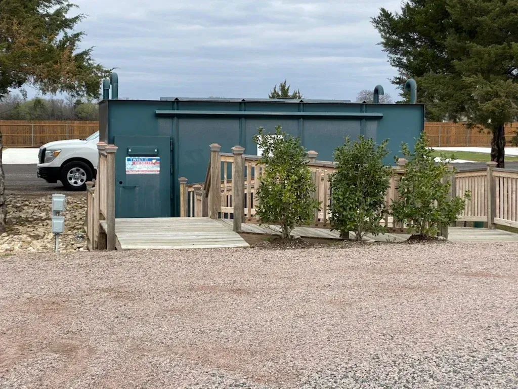 Teal utility building beside a gravel lot, with a wooden ramp and small fenced area.