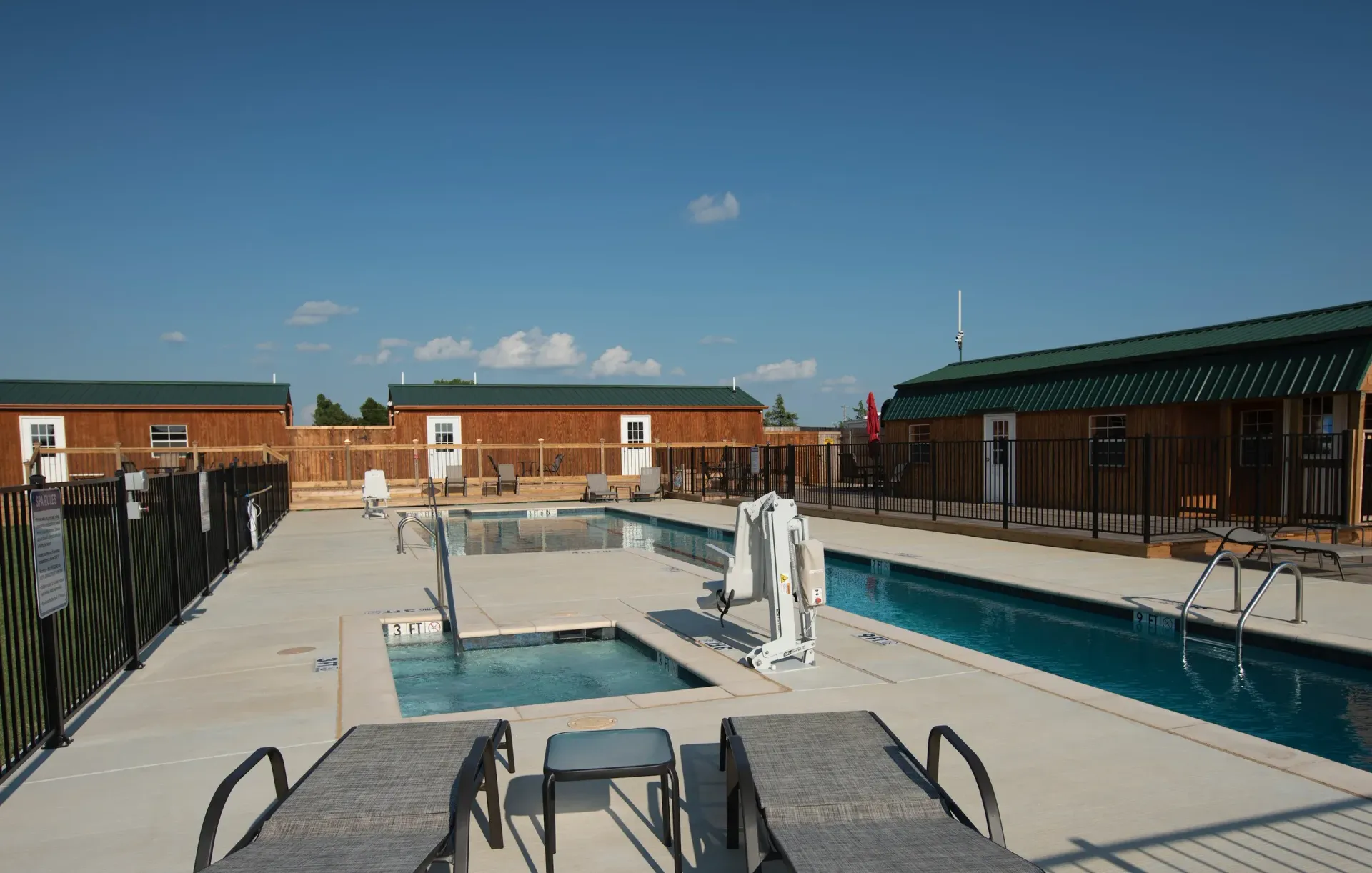 Outdoor swimming pool with lounge chairs, white diving board, and brick buildings under a blue sky