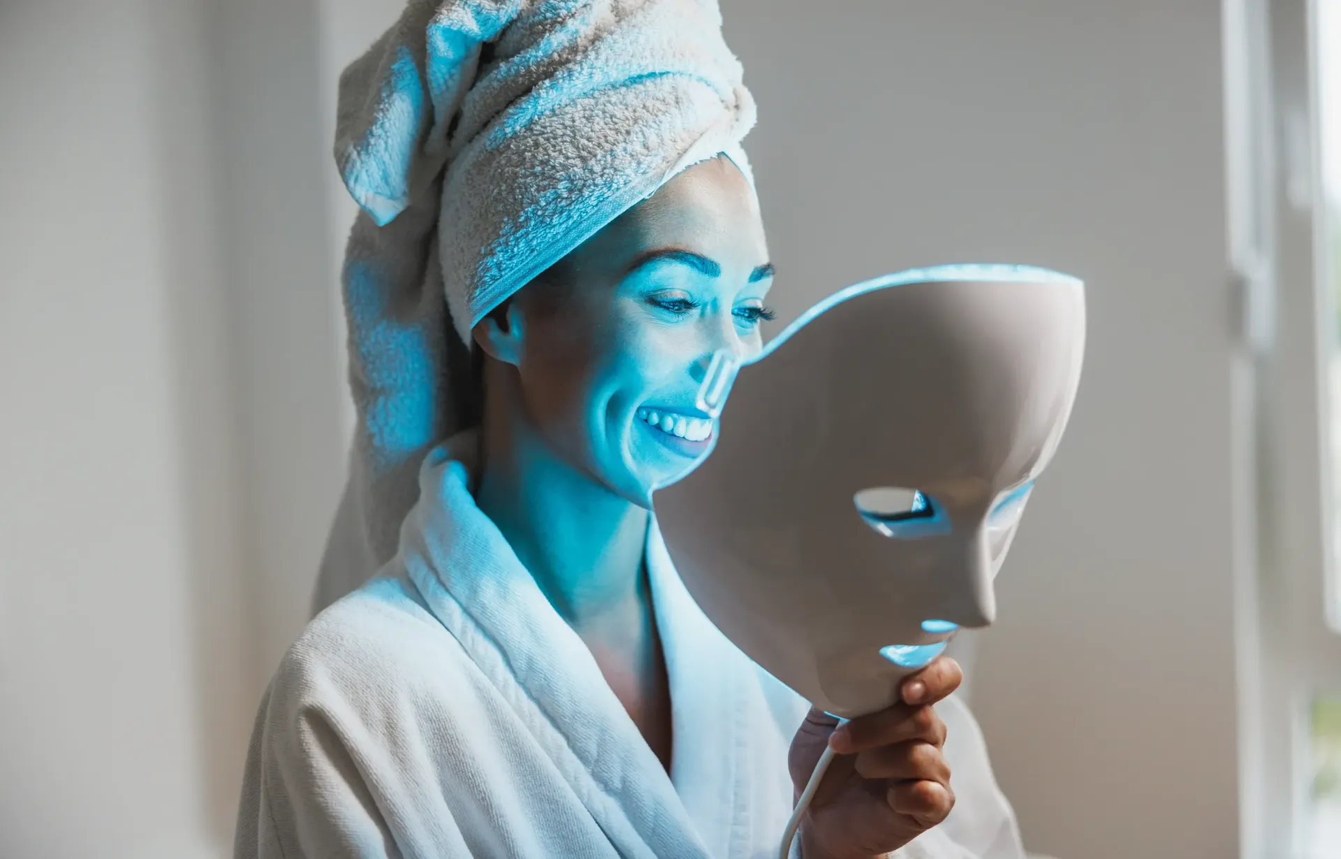 Woman in a robe with a towel on her head holding a facial mask beside a window.