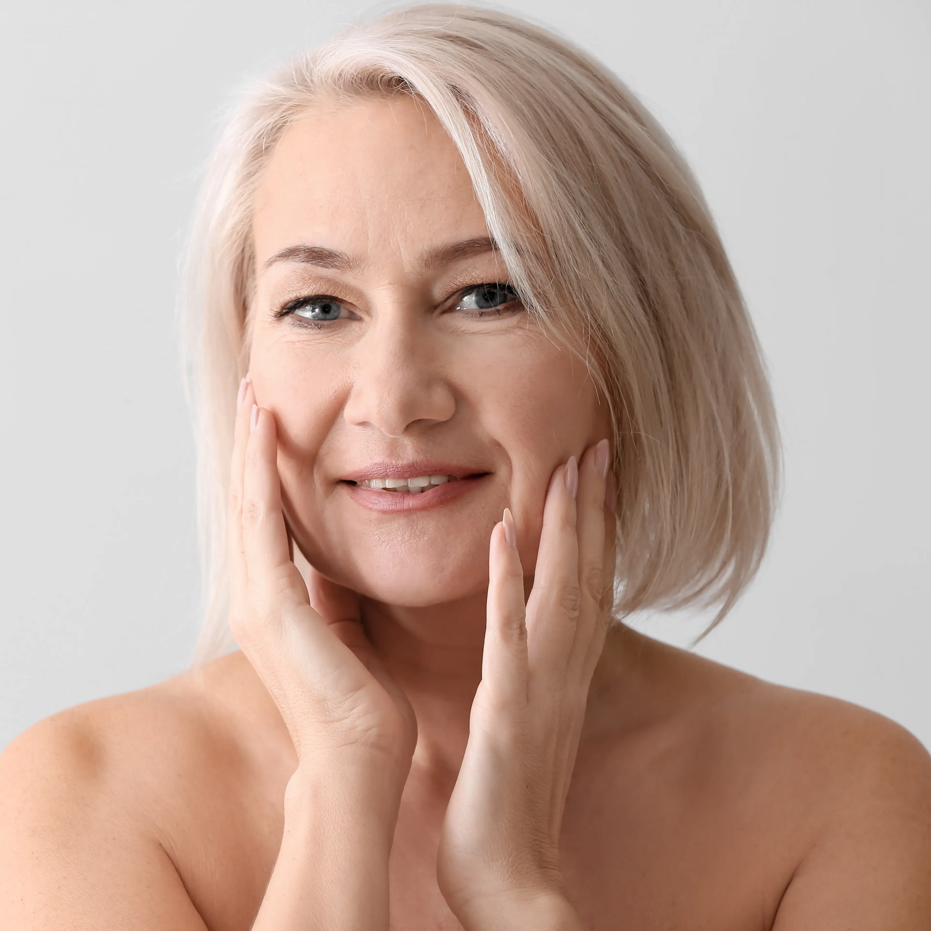 Portrait of a smiling person with short blonde hair, gently touching their cheeks against a light background