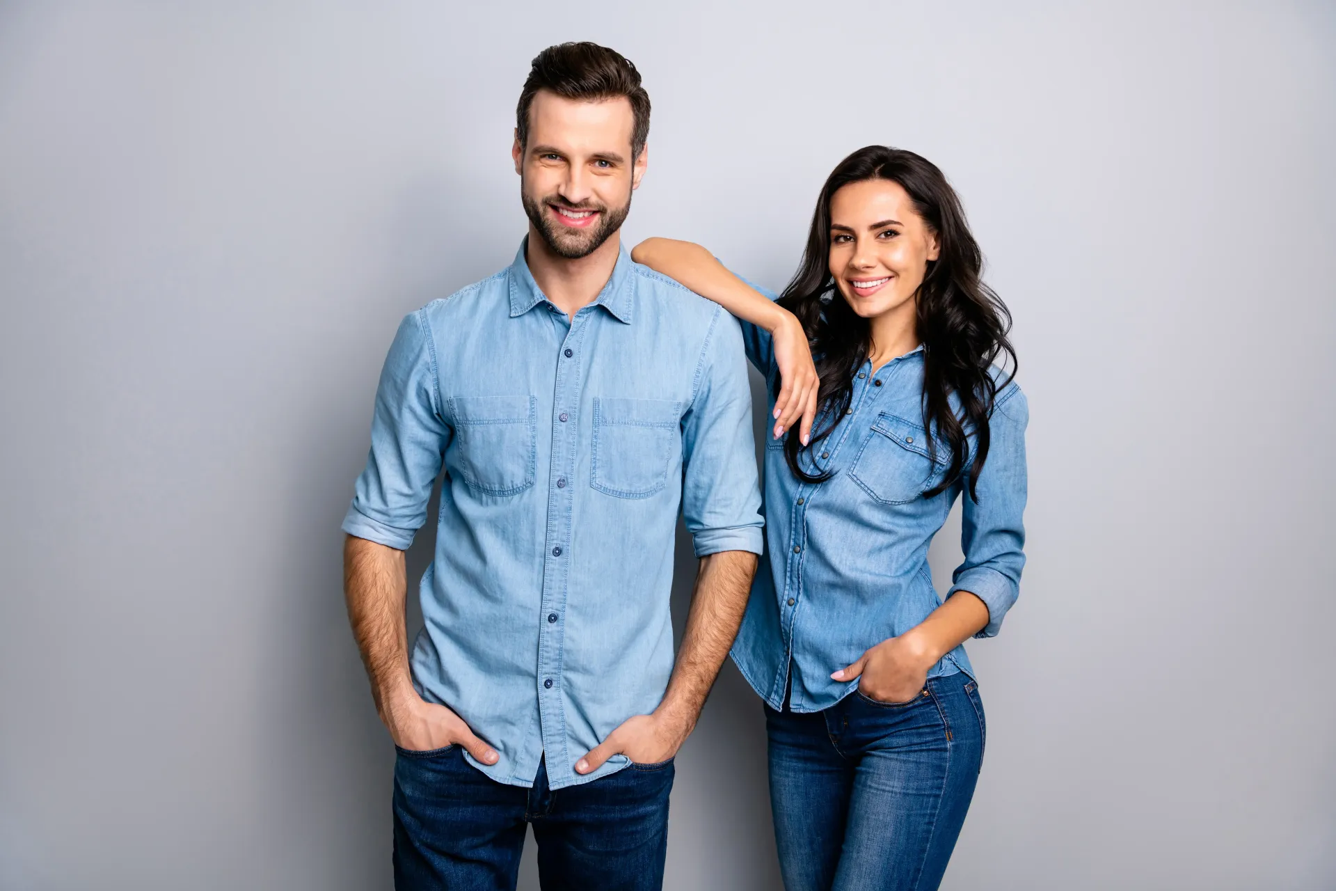Two people in matching denim shirts and jeans posing against a plain gray background.