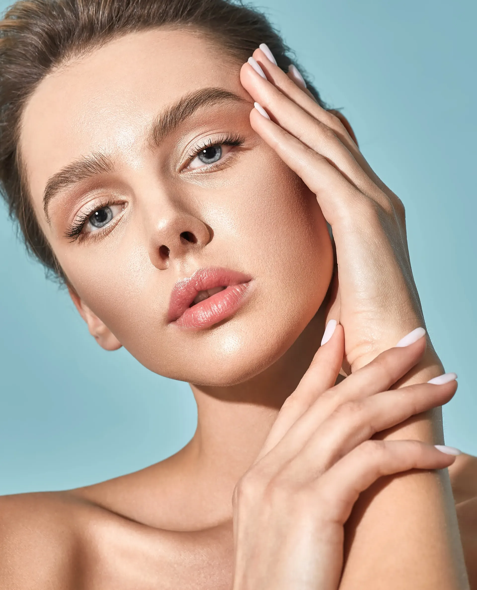 Close-up portrait of a model with glitter makeup, resting hands against her face against a blue background