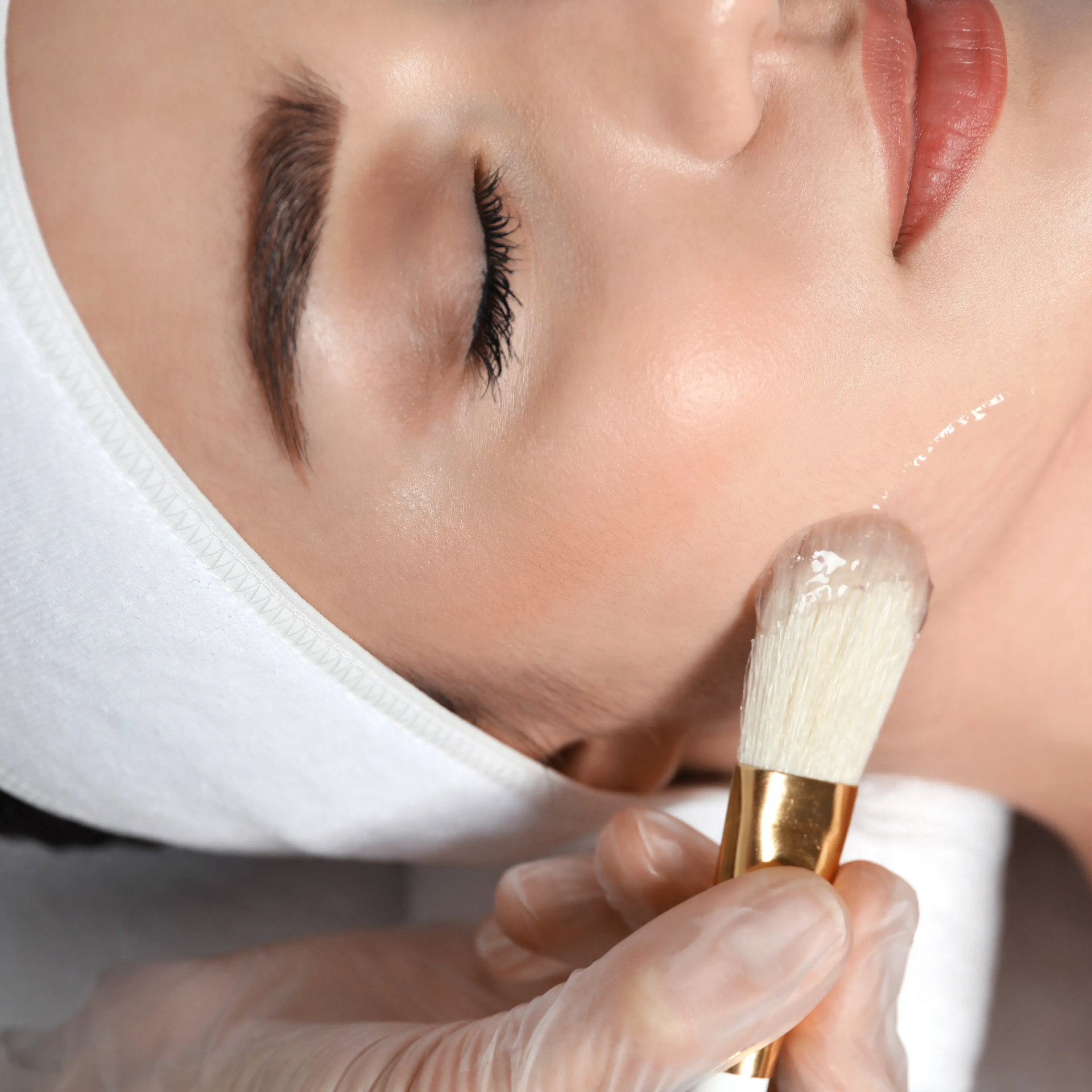 Esthetician applying cream to a woman’s cheek with a brush during a facial treatment