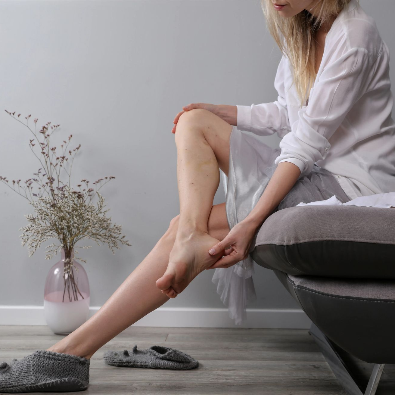 Woman massaging her lower leg while seated on a couch, with slippers on the floor and a vase nearby.