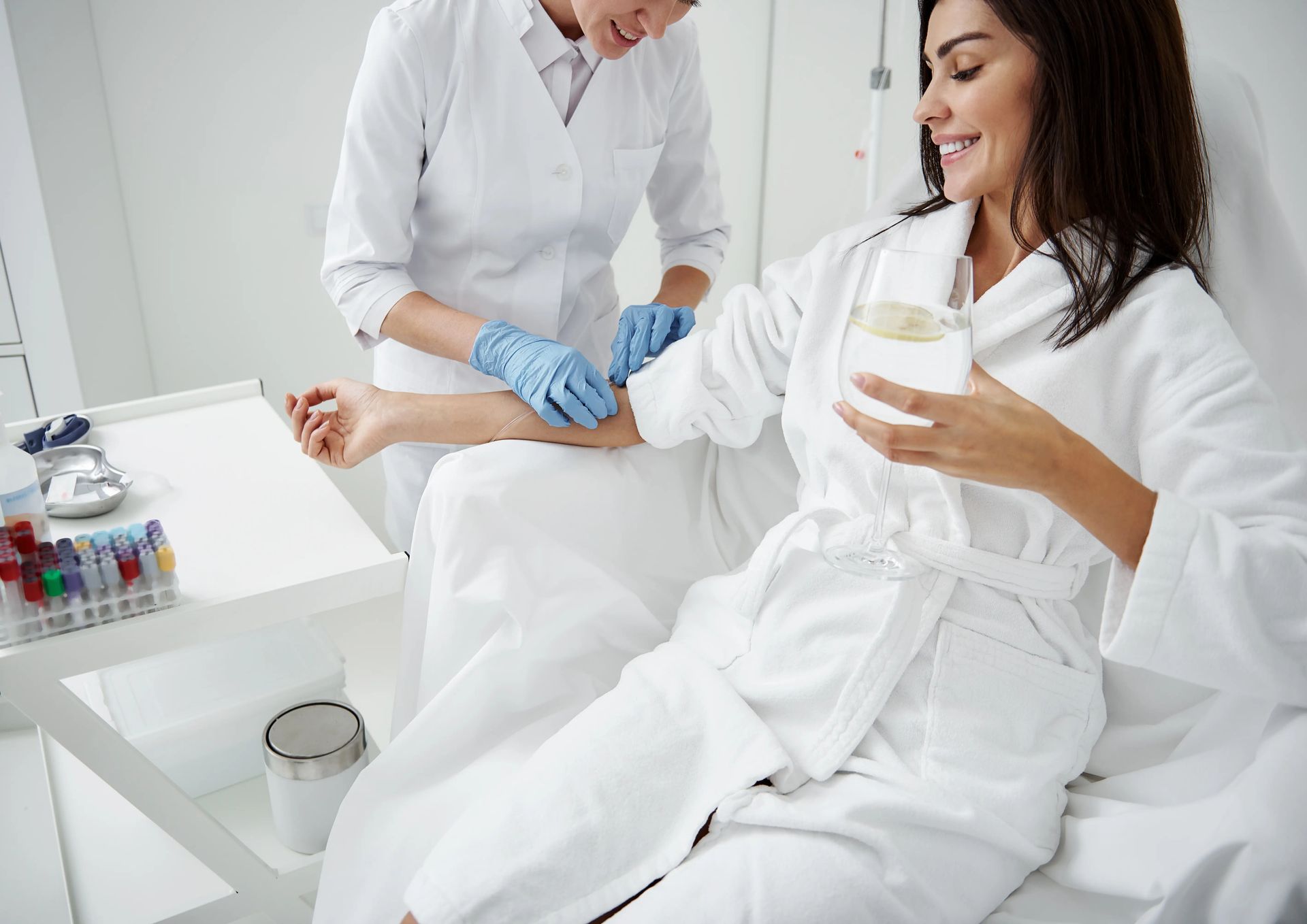 Nurse giving a woman an IV drip while she reclines in a white medical chair holding a drink