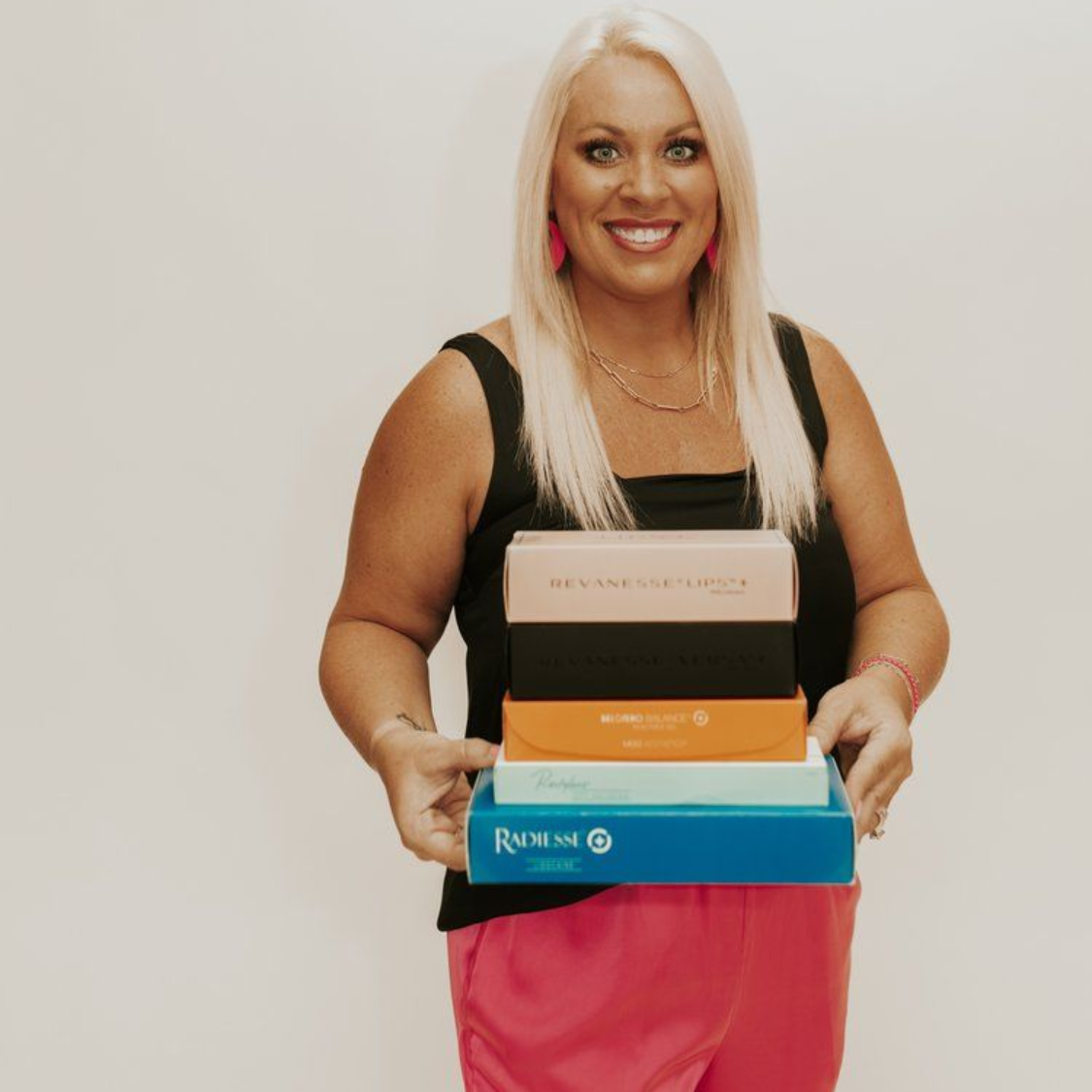 Smiling woman in black top and pink pants holding a stack of colorful boxes against a plain background