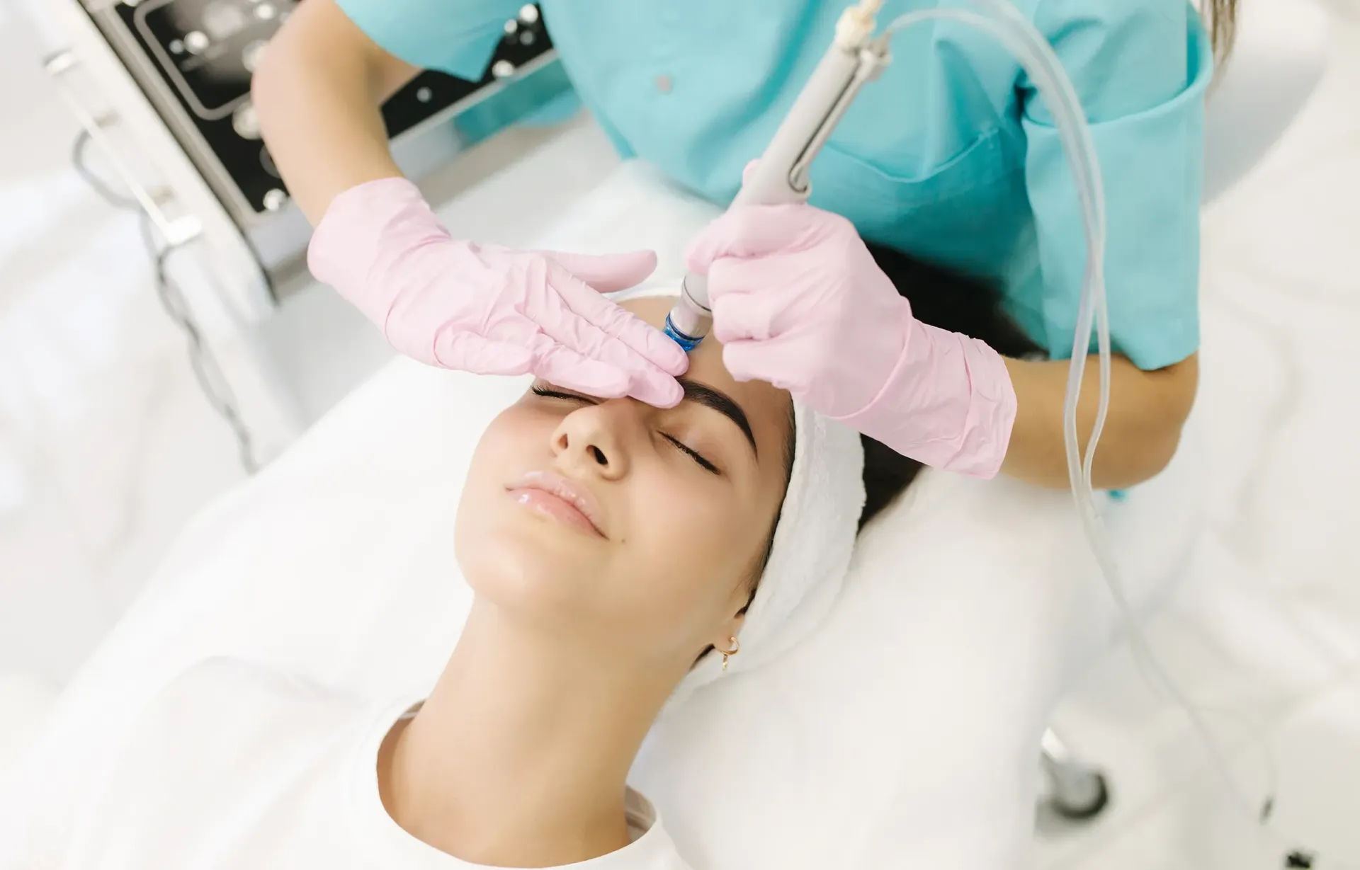 A clinician in pink gloves applies a facial treatment to a patient lying on a treatment bed.