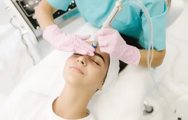 A clinician in pink gloves applies a facial treatment to a patient lying on a treatment bed.