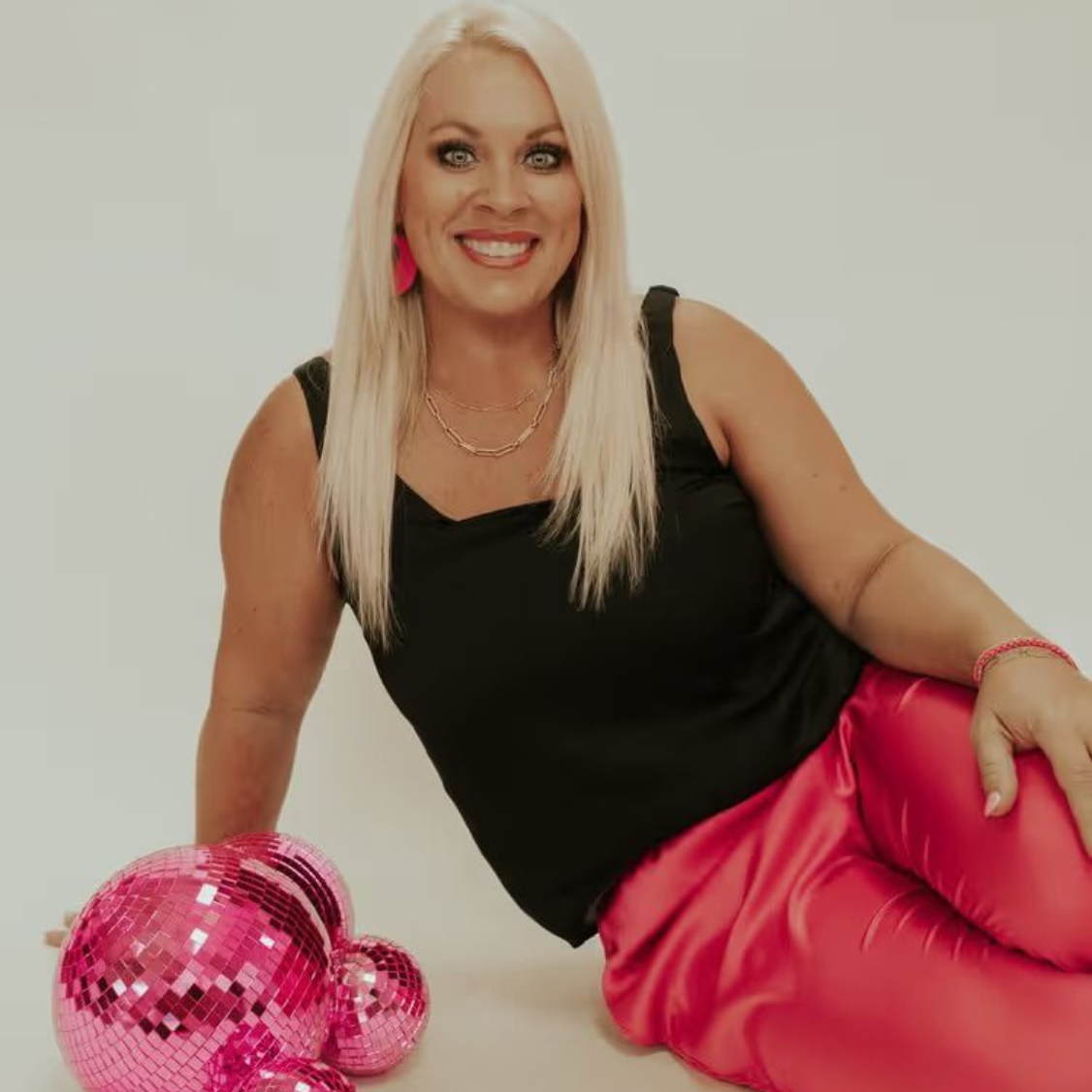 Smiling woman in black top and hot pink pants reclining beside a pink disco ball on a white background