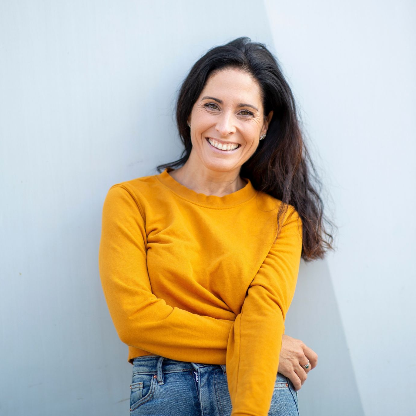 Smiling woman in a mustard sweater and jeans standing against a light blue wall