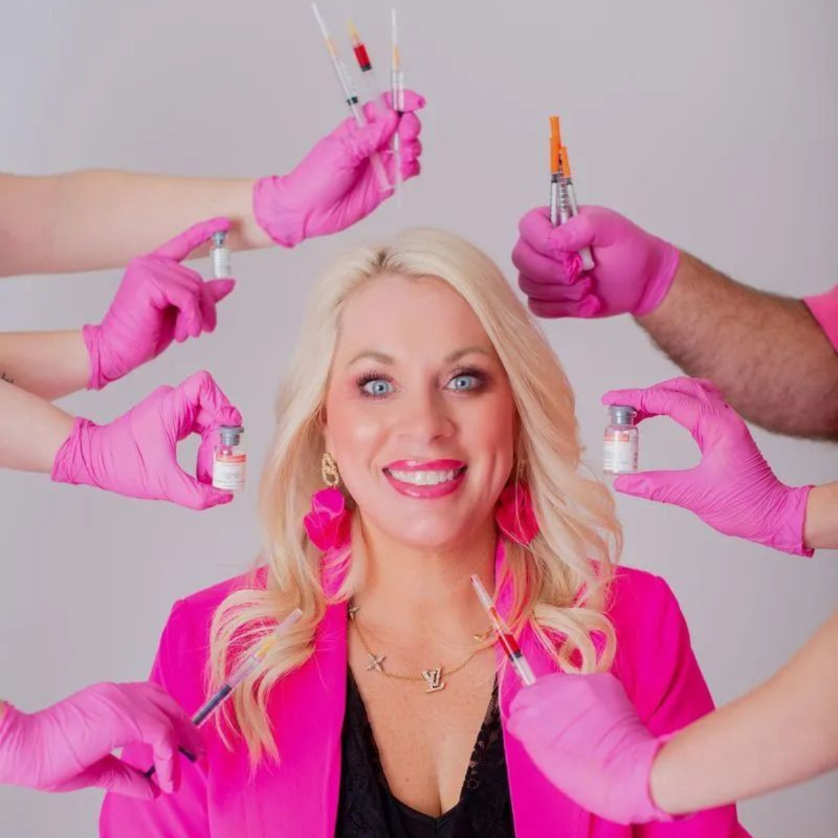 Blonde woman in pink with makeup tools held around her, smiling against a light background