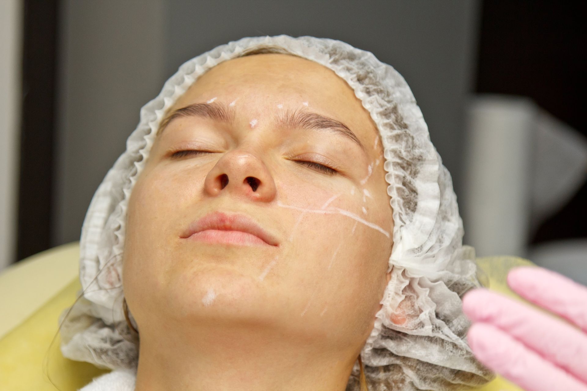 Person with eyes closed wearing a hair cap during a facial treatment, with a gloved hand nearby