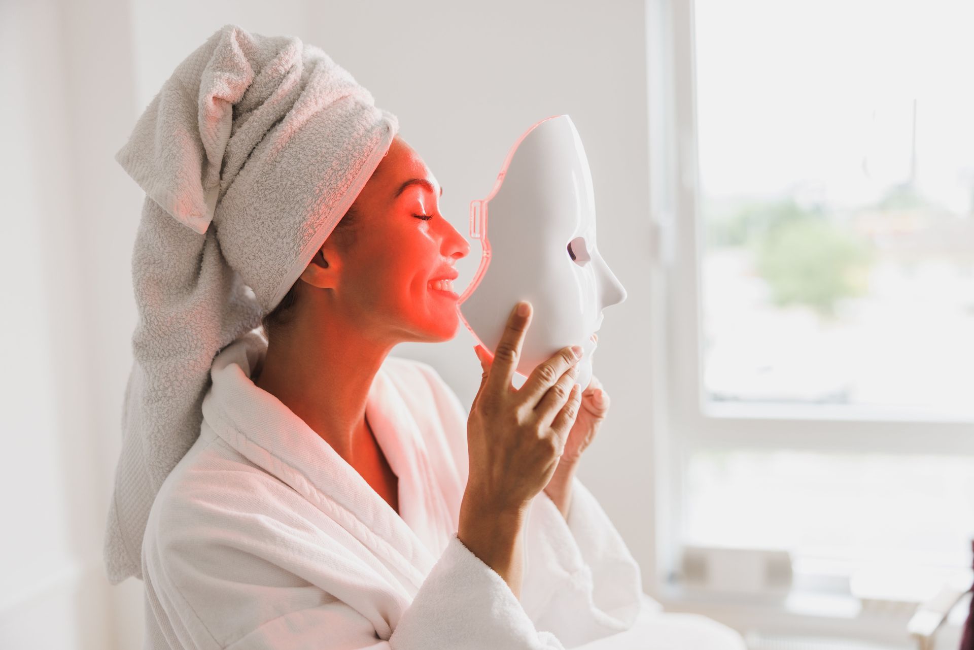 Woman in a white robe using a facial mask beside a bright window
