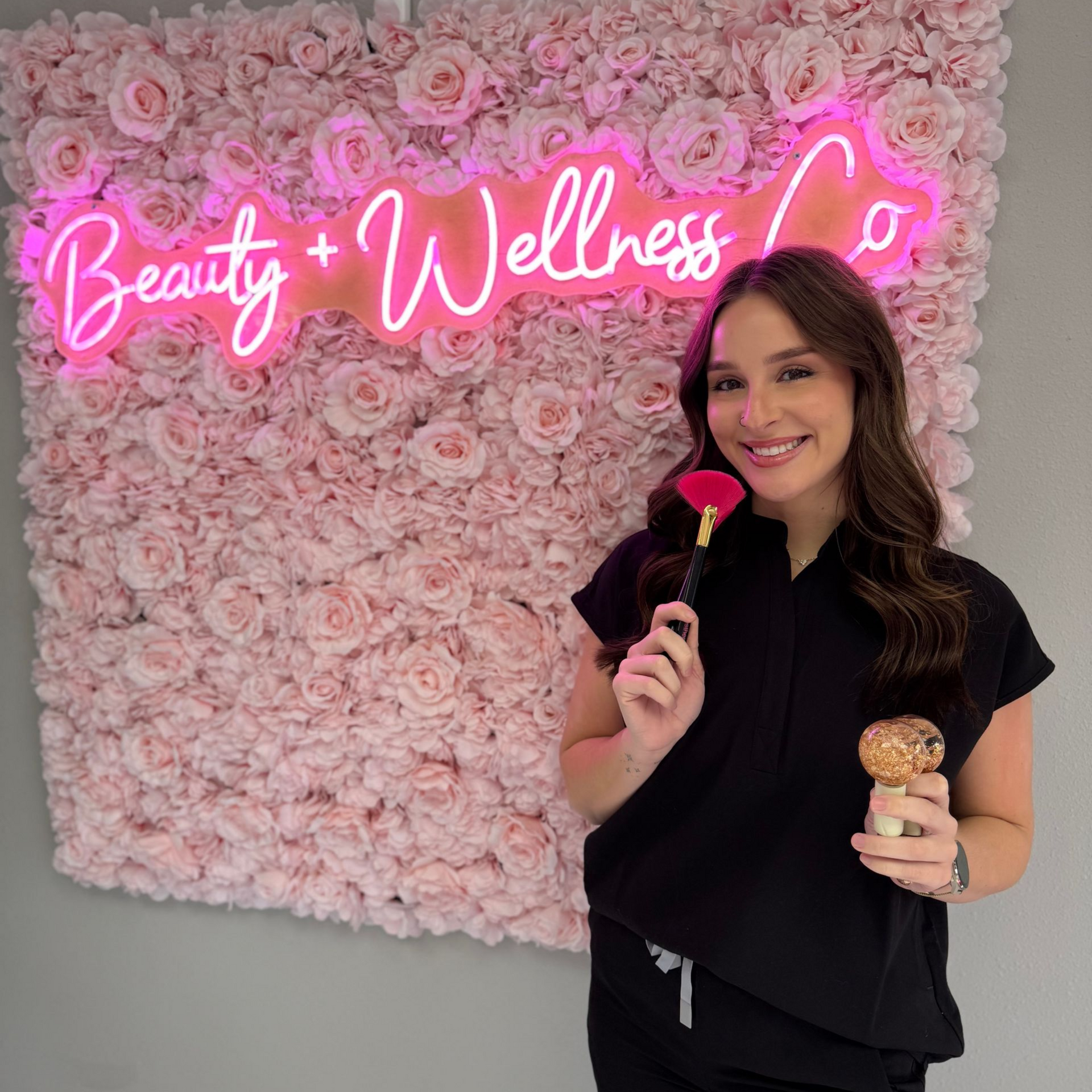 Woman in black holding a pink rose and a treat beside a pink flower wall with neon “Beauty + Wellness” sign