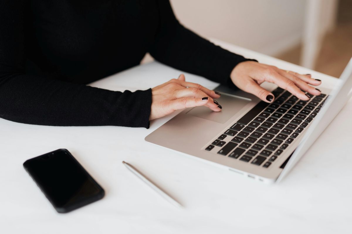A woman is typing on a laptop next to a cell phone and pen
